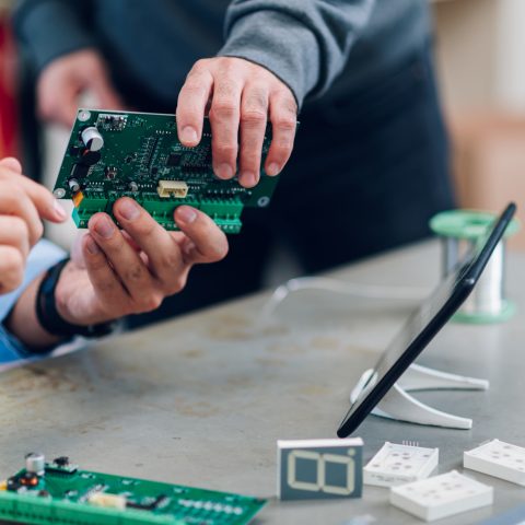 Close shot of a technician hands showing a circuit board to his colleague from work. Electronics engineer working in his workshop. Repairing a device and using a digital tablet. Copy space.